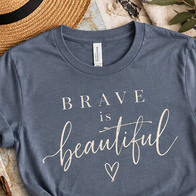 Flat-lay of a slate blue tee printed “Brave is beautiful” in script lettering with a small heart